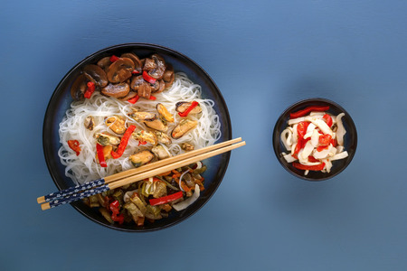 Rice noodles with seafood, salad, red pepper and fried mushrooms in a traditional porcelain plate on a blue table. Top viewの写真素材