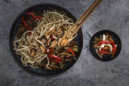 Traditional wooden chopsticks with coiled noodles on the background of a dish with seafood, vegetables and peppers. Close-up. Top viewの写真素材