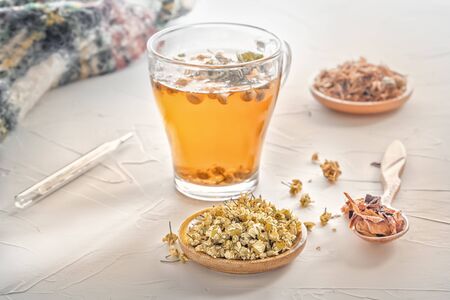Herbal tea with pharmaceutical chamomile, warm scarf and medical thermometer on a white tableの写真素材