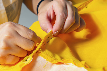 Female hands stitch yellow fabric with a black thread needle in a tailor workshop. Close-up