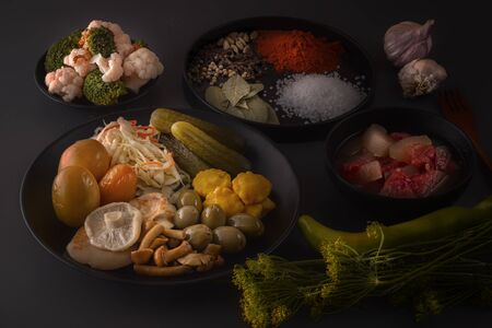 Fermented foods cabbage, peppers, pickles, tomatoes, mushrooms, zucchini, garlic in a black plate on a dark table with spices for salting. Low key. Close-upの写真素材