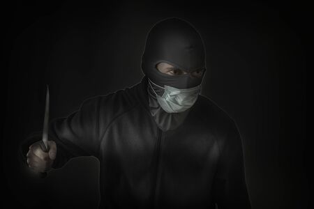 Bandit in black mask with a slit for the eyes and a medical mask waving a knife. Low hatch. Close-up. Copy spaceの写真素材