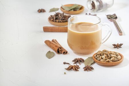 Masala tea in a glass cup and a natural mixture for its preparation - star anise, cinnamon sticks, cardamom, bay leaf, pepper and pepper on a white wooden table. Copy spaceの写真素材