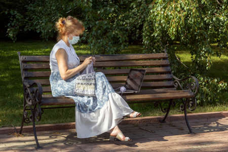 Woman with a medical mask on her face is sitting on a park bench knitting wool clothes on knitting needles and watching lessons or teaching knitting through a laptop. Distance learningの写真素材
