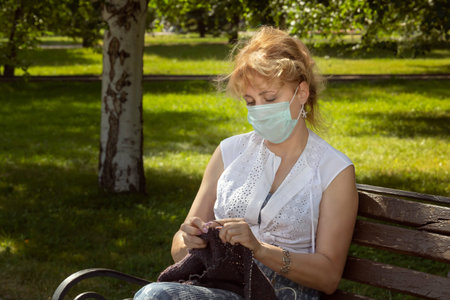 Woman in a medical mask on her face sits on a park bench and knits wool clothes on knitting needles....