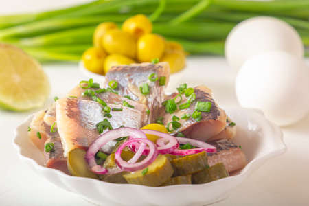 Slices of pickled herring with red onion, fermented cucumber, fried potatoes, olives, boiled eggs and herbs on a light wooden table. Close-upの写真素材
