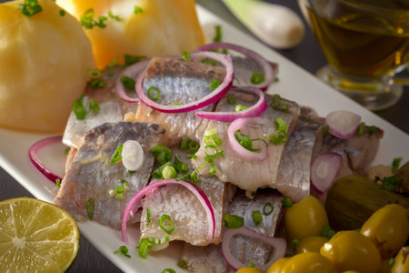 Pickled herring slices with red onion, fermented cucumber, potatoes, olives and lemon in a white...