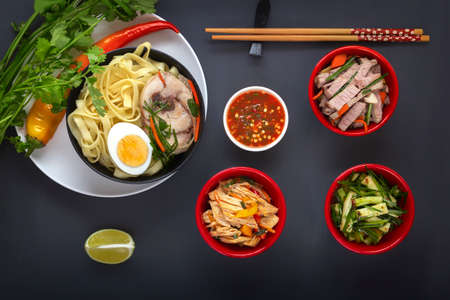 Noodles with pieces of meat and egg in a black plate and dim sums with different traditional snacks of mushrooms, soy and vegetables with chopsticks on a dark table. View from above. Asian food conceptの写真素材
