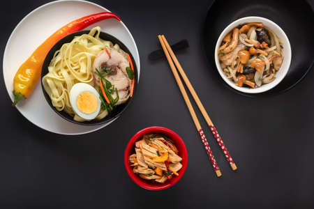 Noodles with pieces of meat and egg in a black plate and dim sums with different traditional snacks of mushrooms, soy and vegetables with chopsticks on a dark table. View from above. Asian food conceptの写真素材