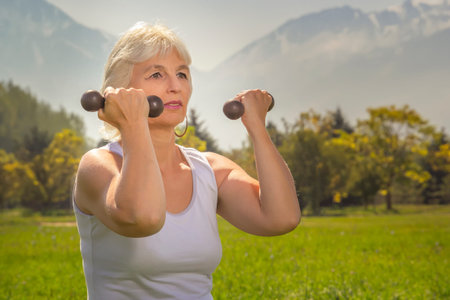 Elderly woman lifts dumbbells while doing fitness in a city park against the backdrop of mountains...