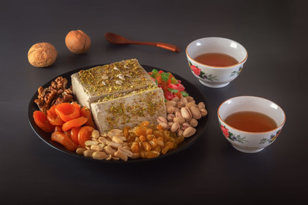 Sunflower halva sprinkled with crushed nuts and raisins on a black plate with pistachios, peanuts,...