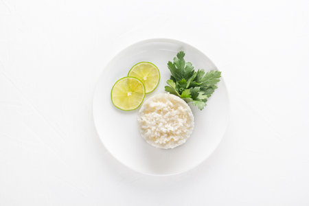 Boiled rice with lemon and parsley leaves in a white plate on a light background. View from above