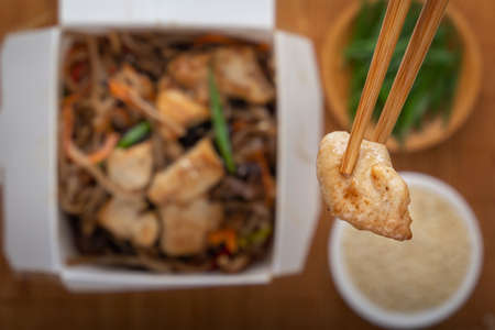 Chopsticks hold a piece of chicken meat over a disposable paper box of dark soba noodles with vegetables, herbs and sesame seeds on a mat background. Asian street food. View from above. Close-upの写真素材