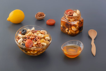 Glass cup and jar filled with a mixture of walnuts, cashews, raisins, dried apricots and prunes with honey, ingredients and lemon on a gray tableの写真素材