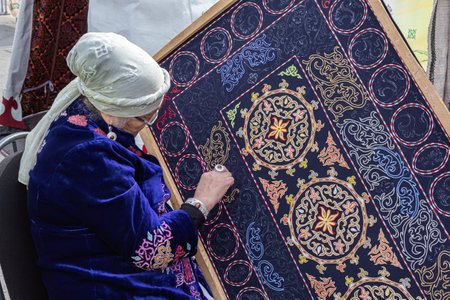 Almaty, Kazakhstan - March 21, 2022. Elderly woman in national clothes embroiders a traditional asian pattern with a thread on a homespun carpetのeditorial素材