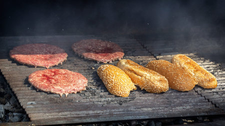 Hamburger patties and bun halves are cooked in smoke on the grill grate of a street fast food...