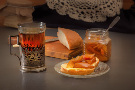 Glass of hot tea in an antique silver cup holder, a piece of rustic bread with pear jam and a jar of jam on a gray wooden table. minimalism. village breakfastの写真素材