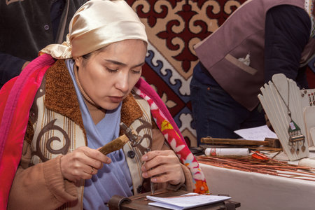 Almaty, Kazakhstan - March 23, 2023. Girl in national dress works a strip of metal for a bracelet with a hammer against the background of the felt walls of a yurt with traditional Asian ornaments. portrait. close-upのeditorial素材