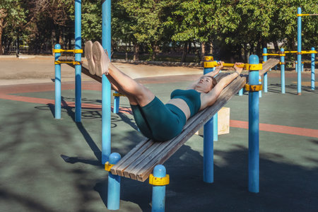 Attractive Caucasian woman performs an abs exercise on a wooden bench at an outdoor sports ground. healthy lifestyleの写真素材