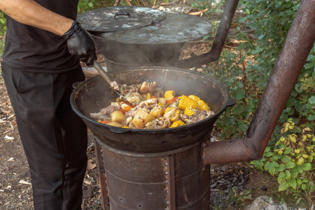 cook uses a slotted spoon to stir meat, corn on the cob, potatoes and vegetables in a cast-iron cauldron on an outdoor wood-burning stove. Street foodの写真素材