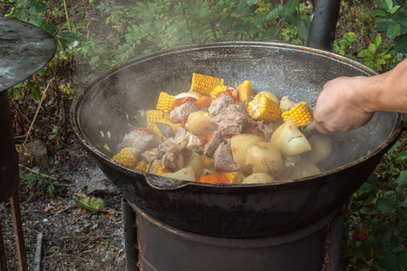 Large pieces of meat, ears of corn, potatoes and vegetables are fried in a cast-iron cauldron on an outdoor wood-burning stove. Street foodの写真素材
