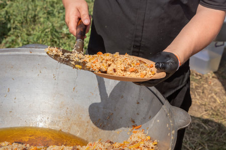 Cook puts pilaf with meat and carrots on a disposable paper plate against the backdrop of a large cast iron cauldron. Street foodの写真素材
