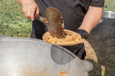 Cook puts pilaf with meat and carrots on a disposable paper plate against the backdrop of a large cast iron cauldron. Street foodの写真素材
