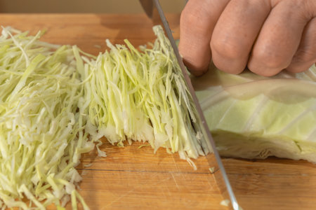 A Cook thinly slices white cabbage with a knife on a wooden cutting board to prepare a vegetable salad. Close-up. Veganism and raw food diet concept.の写真素材