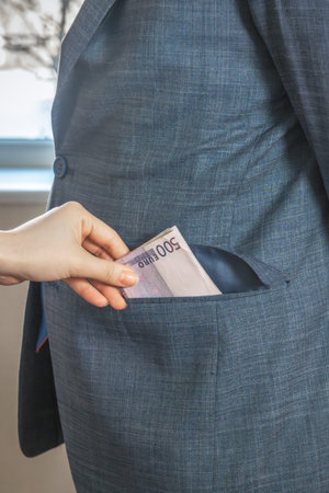 Hand drops European currency banknotes into the pocket of a man in a suit and tie standing in front...