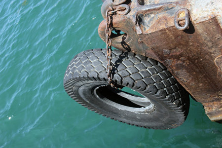 big old black car tire hanging on a chain on the edge of pier as bumper for vesselsの写真素材