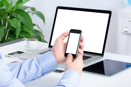Male hands using smartphone mockup with blank screen at the office desk with an open laptop mockup...