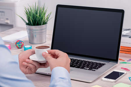 Coffee break with a cup of coffee in mens hands in front of open laptop mockup on office desk. Concept of businessman or student coffee break or rest break at work.の写真素材