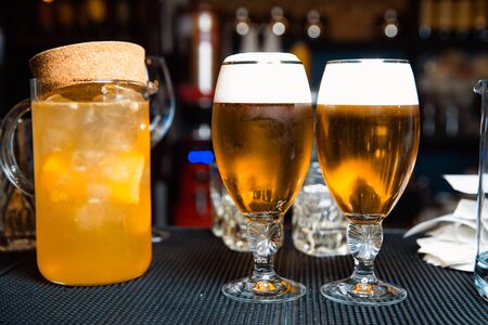 A transparent decanter with orange juice and ice cubes and two glasses of beer is standing at the bar counter.の写真素材