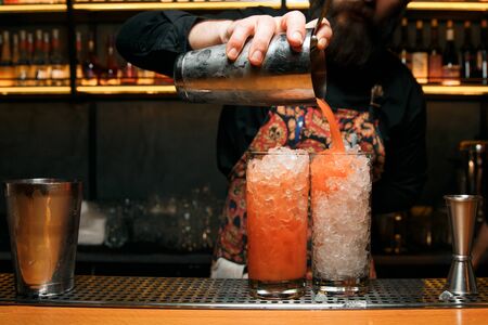 A professional bartender makes two red cocktails, pouring a cocktail from a shaker. Cocktails are on the bar.の写真素材