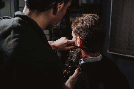 Women professional hairdresser serving client with thick big beard by clipper