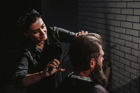Young woman barber making haircut of bearded man in barbershop. Self-care, masculine beautyの写真素材