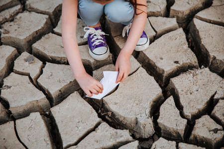 The girl lowers the paper boat onto the dry, cracked ground.の写真素材