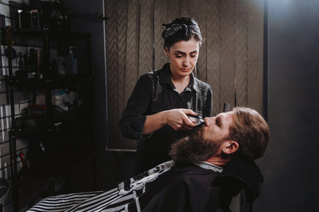Young woman barber making haircut of bearded man in barbershop. Self-care, masculine beautyの写真素材