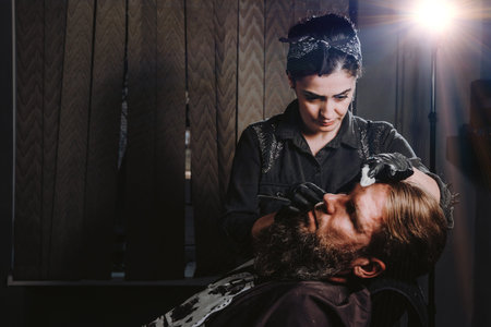 Bearded man getting haircut by hairdresser with dangerous razor at barbershopの写真素材