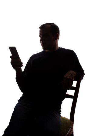 Young man sits in a chair with a mobile phone in his hand - silhouette, Internet concept, communicationの写真素材