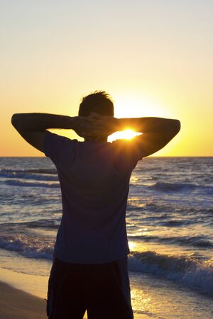 Young man hands behind head relax at sunrise on a background of blue sky and sea, horizon beach dawnの写真素材