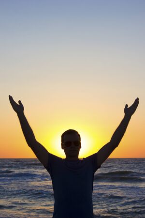 Young man with hands up at sunrise on a background of blue sky and sea, horizon beach dawnの写真素材