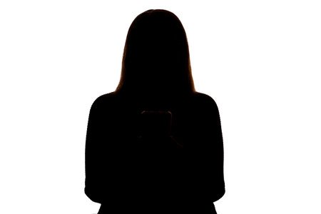 Silhouette of a young woman holding a smartphone and looking at the screen. Isolated on a white background. The scene suggests themes of modern communication, technology, solitude, and personal reflection. Clean and minimalist composition, ideal for concepts related to digital lifestyle, connection, or contemporary identity.の写真素材