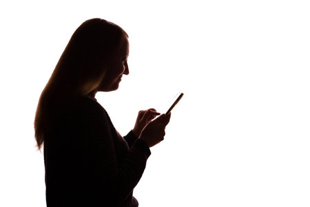 Silhouette of a young woman holding a smartphone and looking at the screen. Isolated on a white background. The scene suggests themes of modern communication, technology, solitude, and personal reflection. Clean and minimalist composition, ideal for concepts related to digital lifestyle, connection, or contemporary identity.の写真素材
