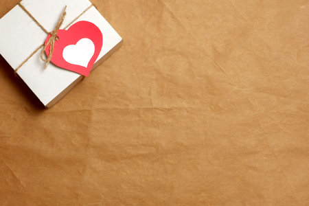White gift box wrapped in brown craft paper, tied with twine with a bow and label in a red heart form on craft paper background. Love, Valentine's, mother's, women's day, relations, romantic conceptの写真素材
