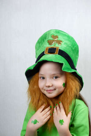 Saint Patricks Day. Endearing smiling small girl with decorative red beard, green shamrok leaf on her cheek and leprechaun hat, raised her hands with green nails and clover leaves to beardの写真素材