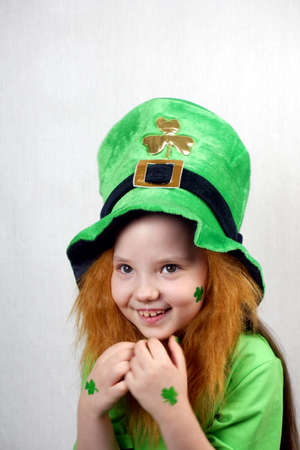 Laughting cute small girl with decorative red beard, green shamrok leaf on her cheek and leprechaun hat, raised her hands with green nails and clover leavesの写真素材