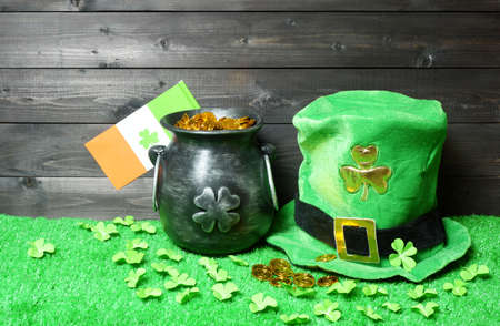 Cast iron pot with treasure, irish flag with clover leaf and leprehaun hat, clover leaves and coins on green grass, dark wooden planks background. Saint Patricks Day banner, poster, flyer, invitationの写真素材
