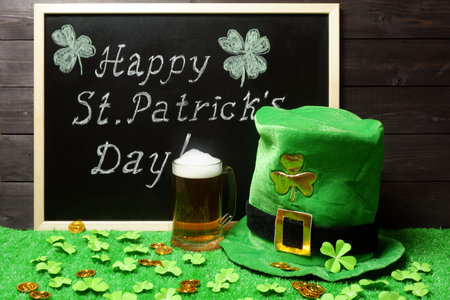Happy St. Patricks Day chalk lettering on blackboard, mug of beer, leprechauns hat and shamrock leaves and coins on green grass, dark wooden planks background. Saint Patricks Day templateの写真素材