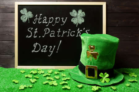 Happy St. Patricks Day chalk lettering on blackboard, green leprechaun hat and shamrock leaves on green grass. Dark wooden planks background. Saint Patricks Day banner, poster, invitation templateの写真素材